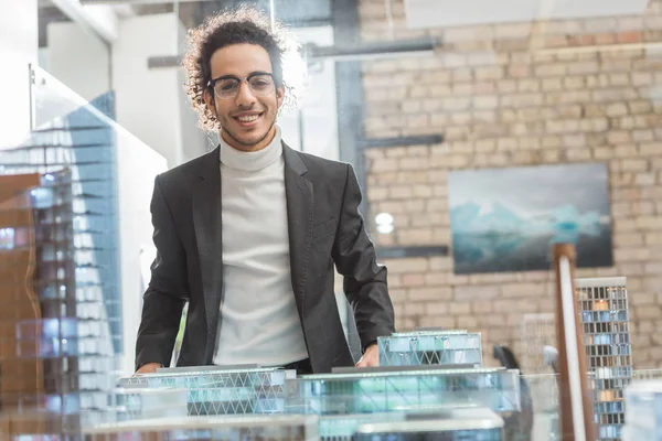 Glücklicher junger Architekt im Anzug in der Nähe von Gebäuden Miniaturmodell im Büro — Stockfoto
