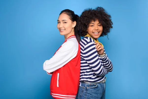 Smiling african american and asian girls standing back to back, isolated on blue — Stock Photo