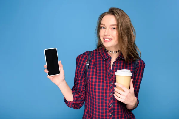 Hermosa joven sosteniendo la taza en el café y mostrando teléfono inteligente, aislado en azul — Stock Photo