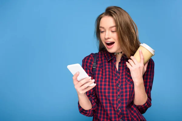 Chica sorprendida con café para ir con teléfono inteligente, aislado en azul - foto de stock