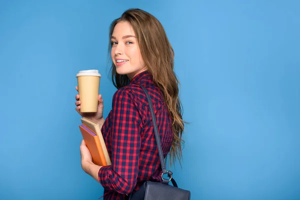 Joven estudiante sonriente de pie con libros, mochila y café para llevar, aislado en azul - foto de stock