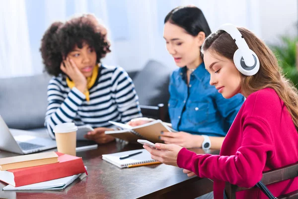 Enfoque selectivo de los jóvenes estudiantes en los auriculares utilizando el teléfono inteligente con amigos multiétnicos cerca - foto de stock