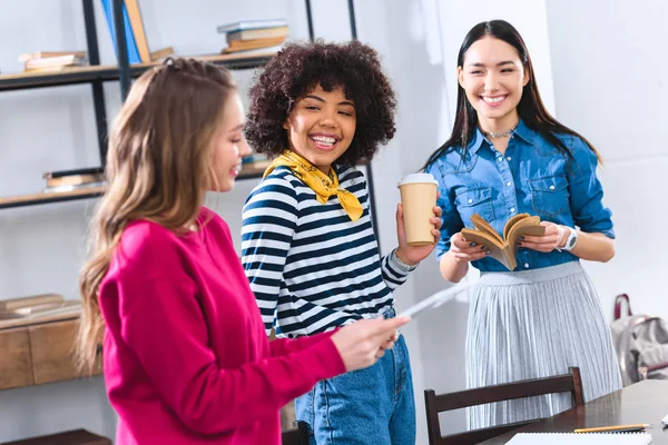 Selective focus of smiling multicultural students with tablet, book and coffee to go — Stock Photo