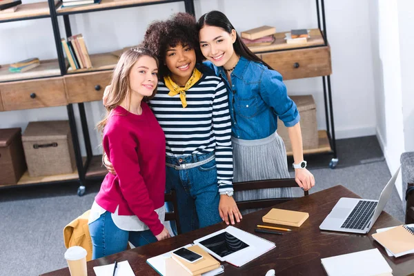 Vista ad alto angolo di studenti multiculturali sorridenti in piedi a tavola con quaderni e dispositivi digitali — Foto stock