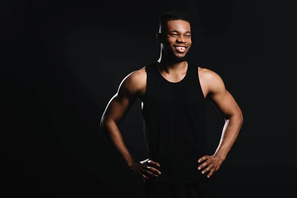 Smiling muscular african american man standing with hands on waist and looking away isolated on black — Stock Photo