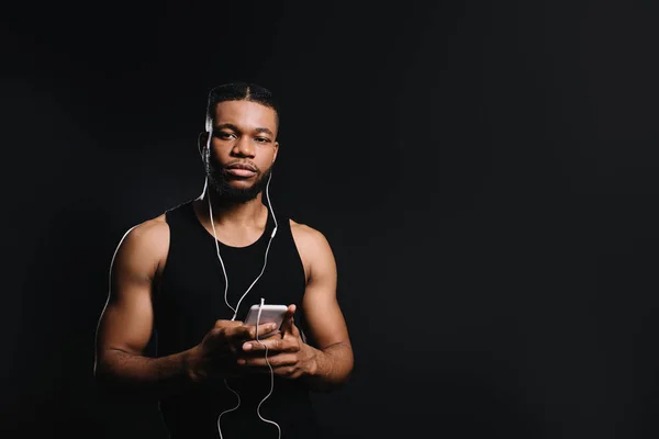 Young african american sportsman in earphones holding smartphone and looking at camera isolated on black — Stock Photo