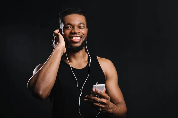 Hombre americano africano muscular en auriculares usando teléfono inteligente y sonriendo a la cámara aislada en negro - foto de stock