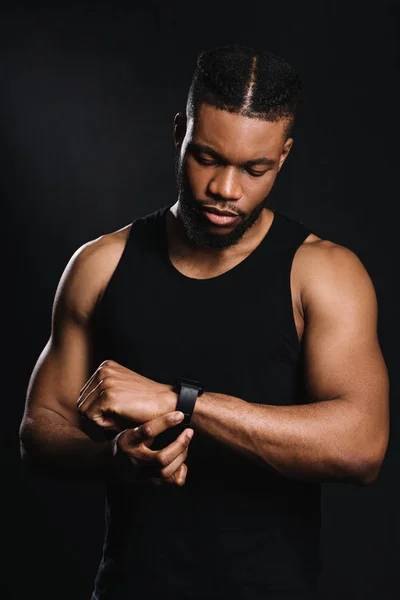 Guapo serio afroamericano deportista usando smartwatch aislado en negro - foto de stock