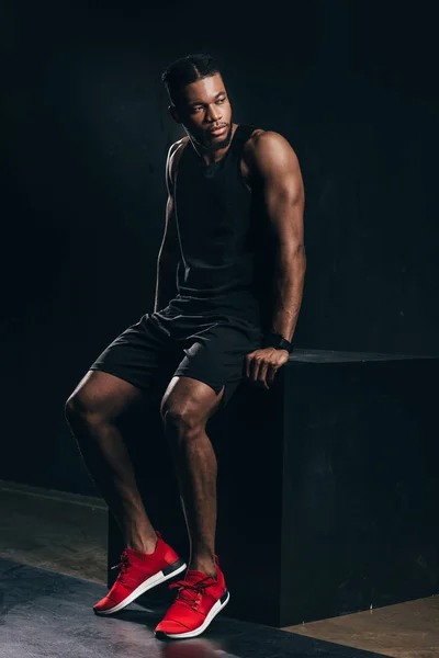 Handsome young african american sportsman sitting and looking away on black — Stock Photo