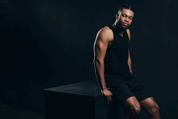 Handsome young african american sportsman sitting and looking at camera on black — Stock Photo