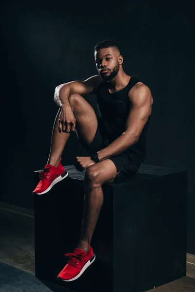 Vue pleine longueur de musclé jeune homme afro-américain en vêtements de sport assis sur noir — Photo de stock