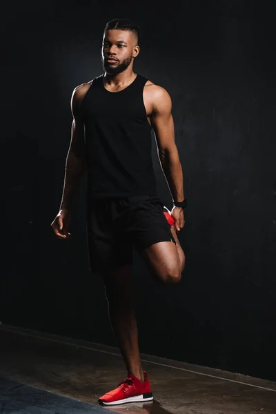 Vista completa del joven afroamericano deportista estirando los músculos y mirando hacia otro lado en negro - foto de stock