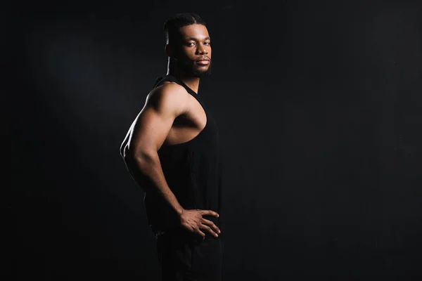 Вид сбоку атлетичный молодой африканский американец, стоящий с руками на талии и смотрящий на камеру, изолированную на черном — стоковое фото