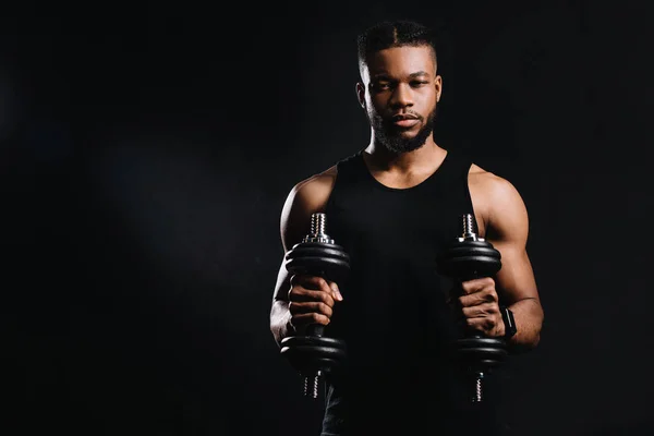 Musculoso joven afroamericano deportista sosteniendo mancuernas y mirando a la cámara aislado en negro - foto de stock