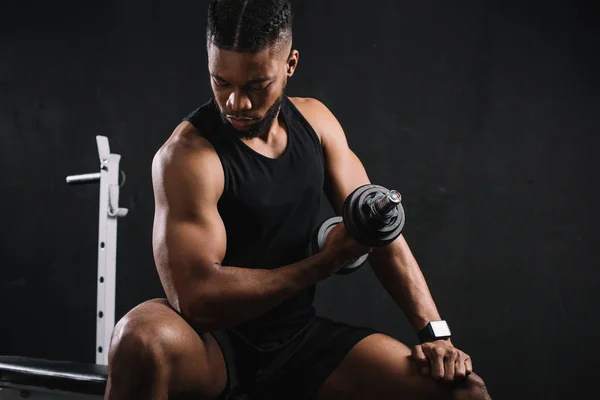 Joven deportista afroamericano ejercitándose con mancuerna y mirando bíceps - foto de stock