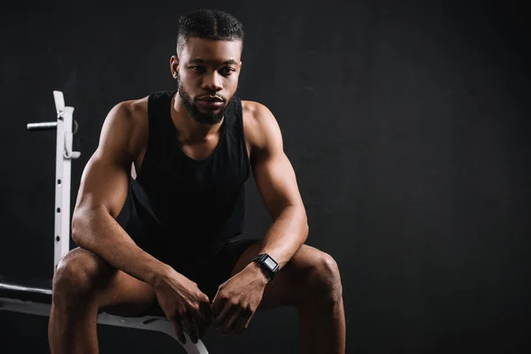 Musclé jeune homme afro-américain en vêtements de sport assis sur noir — Photo de stock