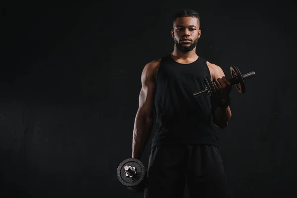 Joven deportista afroamericano haciendo ejercicio con mancuernas y mirando a la cámara aislada en negro - foto de stock