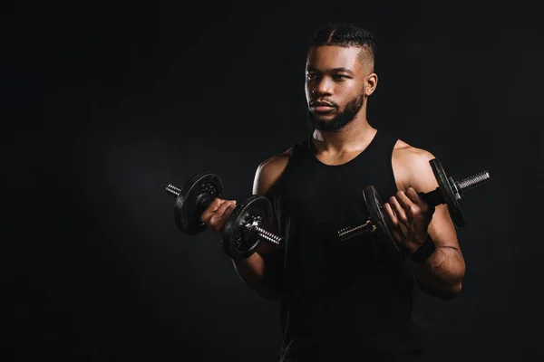 Joven deportista afroamericano ejercitándose con mancuernas y mirando hacia otro lado aislado en negro - foto de stock