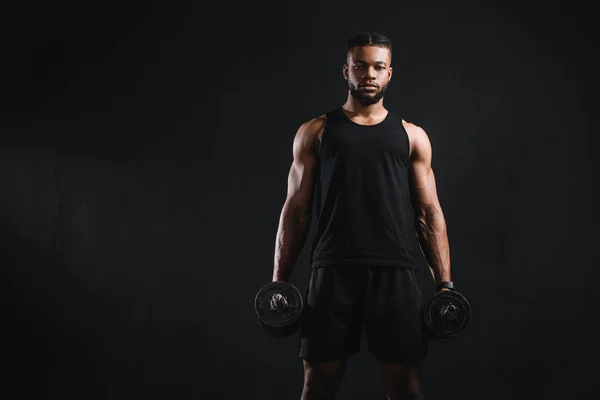 Joven musculoso afroamericano deportista sosteniendo mancuernas y mirando a la cámara aislado en negro - foto de stock