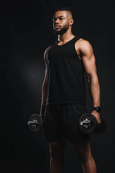 Musculoso joven afroamericano hombre sosteniendo mancuernas y mirando hacia otro lado aislado en negro - foto de stock