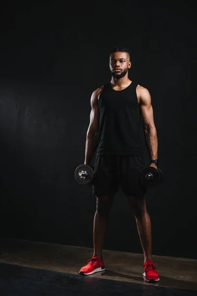 Vista completa de joven muscular afroamericano deportista sosteniendo mancuernas en negro - foto de stock