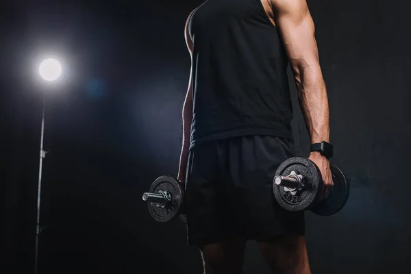 Cropped shot of young african american sportsman holding dumbbells on black — Stock Photo