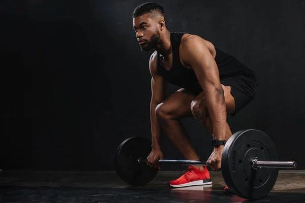 Muscular young african american sportsman lifting barbell and looking away on black — Stock Photo