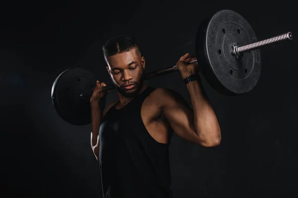 Musclé jeune sportif afro-américain levant haltère et regardant biceps sur noir — Photo de stock