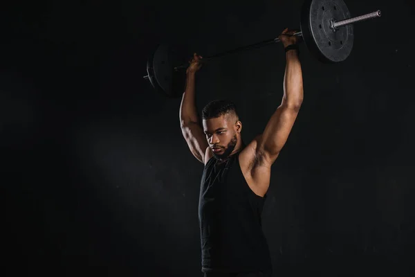 Musculoso joven afroamericano deportista levantando la barra y mirando hacia otro lado aislado en negro - foto de stock