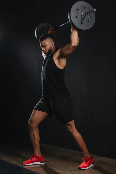 Vista completa de la musculosa joven afroamericano deportista levantando la barra en negro - foto de stock