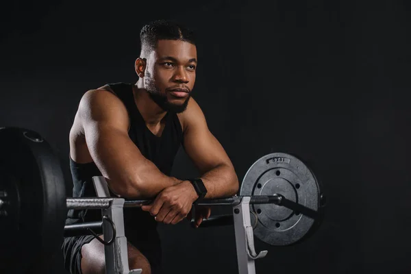 Jovem desportista afro-americano muscular inclinando-se em barbell e olhando para longe em preto — Fotografia de Stock