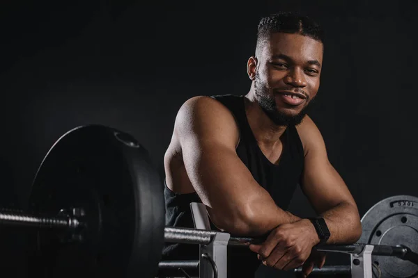 Muscular young african american sportsman leaning at barbell and smiling at camera on black — Stock Photo