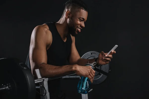 Sonriente joven afroamericano deportista sosteniendo la botella de agua y el uso de teléfono inteligente - foto de stock