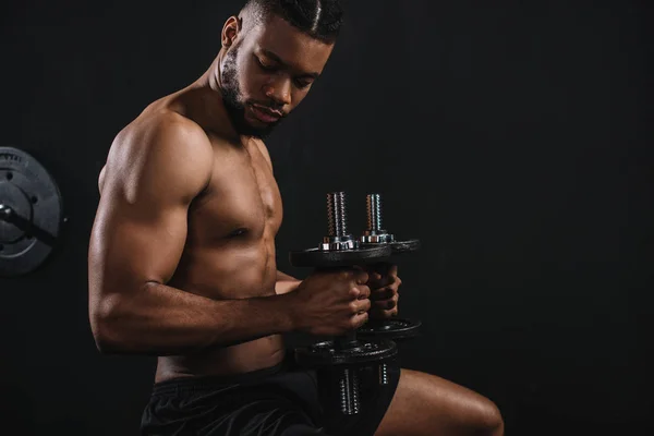 Vista lateral de musculoso sin camisa joven afroamericano hombre ejercitando con mancuernas en negro - foto de stock