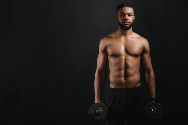 Muscular sem camisa afro-americano homem segurando halteres e olhando para a câmera isolada no preto — Fotografia de Stock