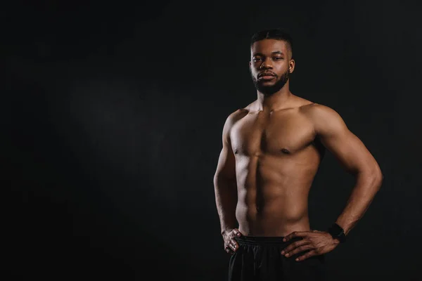 Musclé torse nu afro-américain homme debout avec les mains sur la taille et en regardant la caméra isolée sur noir — Photo de stock