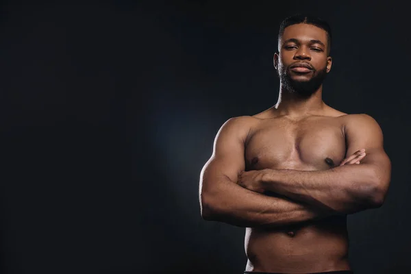 Muskulöser hemdloser junger afrikanisch-amerikanischer Mann, der mit verschränkten Armen steht und isoliert auf schwarz in die Kamera schaut — Stockfoto