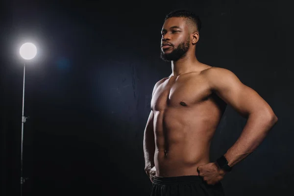 Musclé jeune sportif afro-américain debout avec les mains sur la taille et regardant loin sur noir — Photo de stock