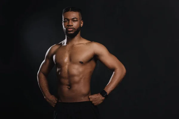 Muskulöser junger afrikanisch-amerikanischer Sportler, der mit den Händen auf der Taille steht und isoliert auf schwarz in die Kamera schaut — Stockfoto