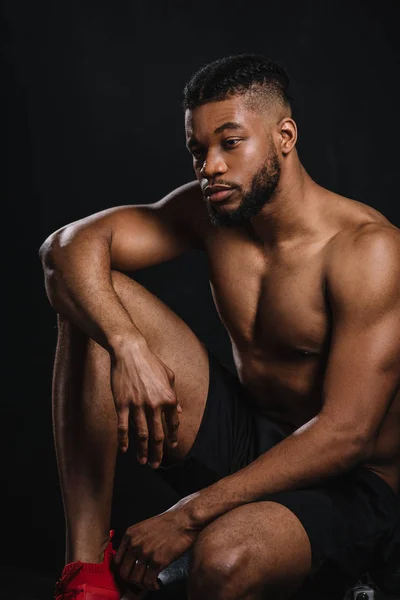 Pensativo sin camisa joven afroamericano deportista sentado y mirando hacia otro lado en negro — Stock Photo