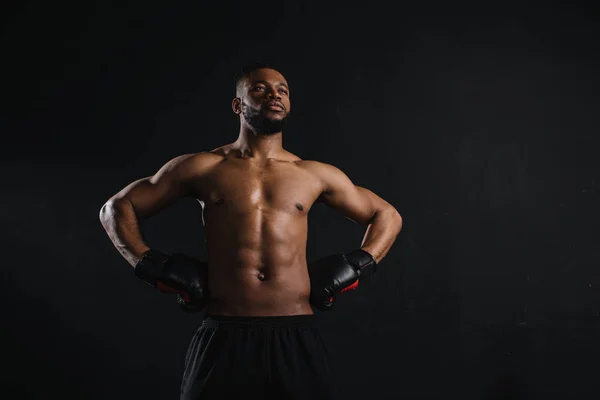 Muscular sem camisa afro-americano homem em luvas de boxe de pé com as mãos na cintura e olhando para longe isolado em preto — Fotografia de Stock