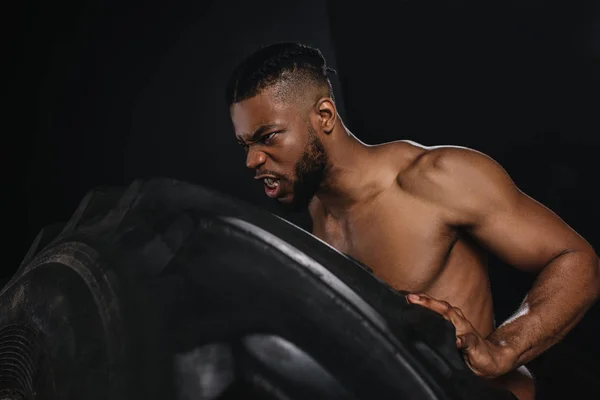 Side view of muscular young african american sportsman lifting tire on black — Stock Photo