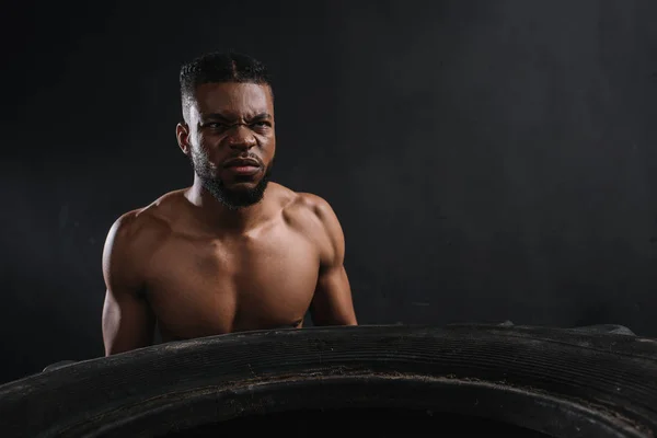 Muscular young african american sportsman lifting tire on black — Stock Photo