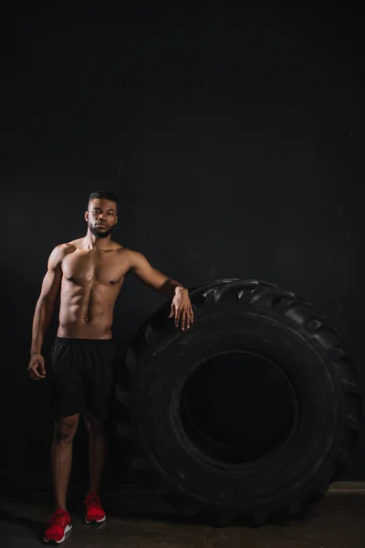 Muscular shirtless young african american sportsman leaning at tyre and looking at camera on black — Stock Photo