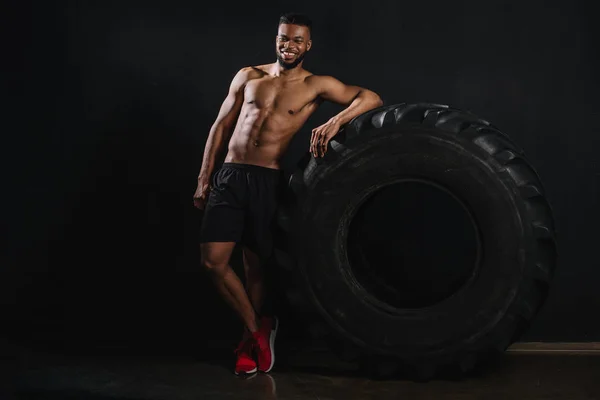 Musculoso sin camisa joven afroamericano deportista apoyado en el neumático y sonriendo a la cámara en negro - foto de stock