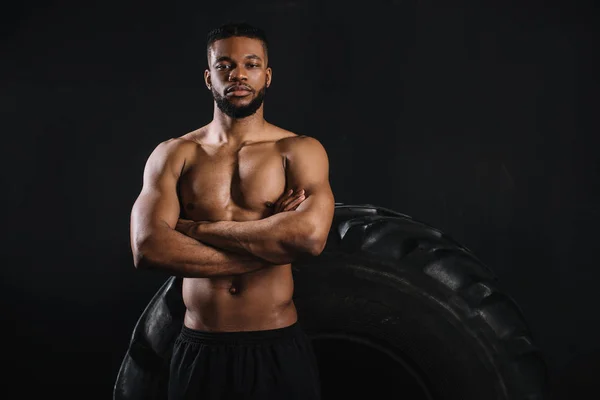 Musculoso deportista afroamericano sin camisa de pie con los brazos cruzados cerca de neumático grande y mirando a la cámara aislada en negro - foto de stock
