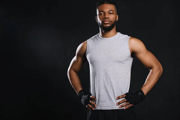 Guapo joven afroamericano deportista de pie con las manos en la cintura y mirando a la cámara aislado en negro - foto de stock