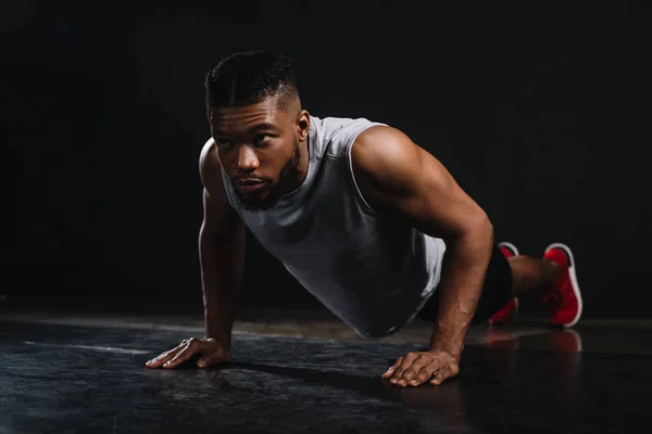 Joven musculoso afroamericano hombre en ropa deportiva haciendo flexiones y mirando hacia otro lado en negro - foto de stock