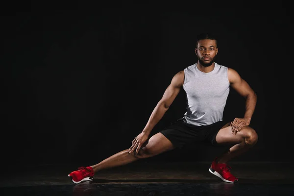 Athletic young african american man in sportswear exercising and looking at camera on black — Stock Photo
