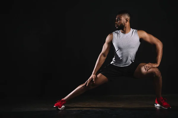 Atlético joven afroamericano hombre en ropa deportiva ejercicio y mirando hacia otro lado en negro - foto de stock